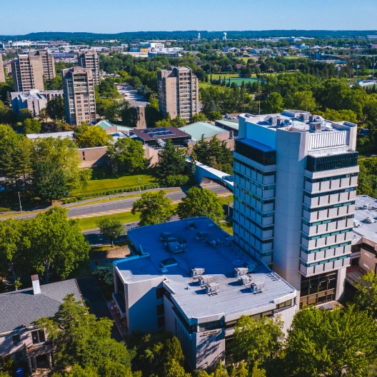 Aerial view of a university campus with tall academic buildings surrounded by trees, roads, and green spaces under a clear blue sky. Residential and academic structures are visible in the background.