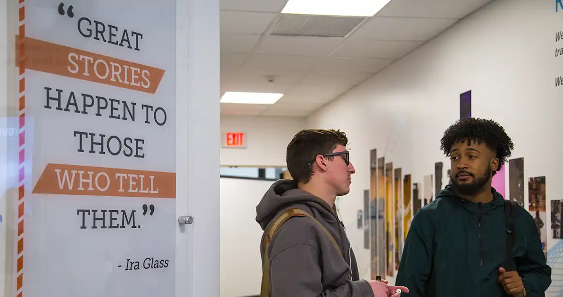 Two students talking to each other in front of a sign that reads "Great stories happen to those who tell them"